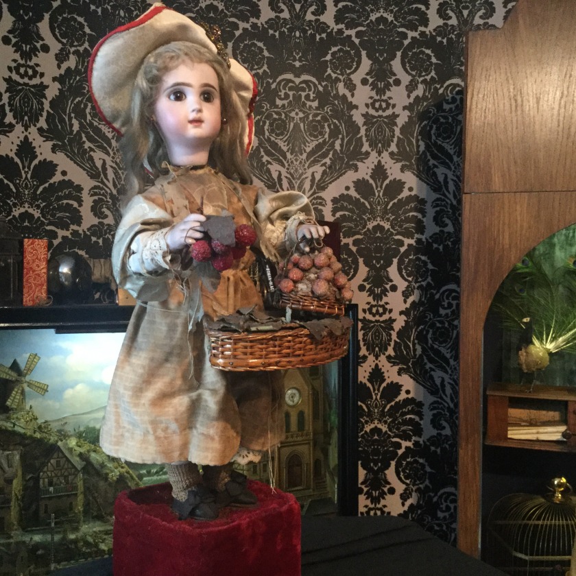 A 'Strawberry Girl' automata, resembling a ceramic doll holding a basket of strawberries, in front of a patterned wallpaper and Victorian diorama.